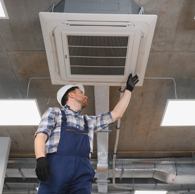 Maintenance climatisation : intervention d'un technicien Technicien inspectant une climatisation cassette au plafond d'un local professionnel avec gants et casque.